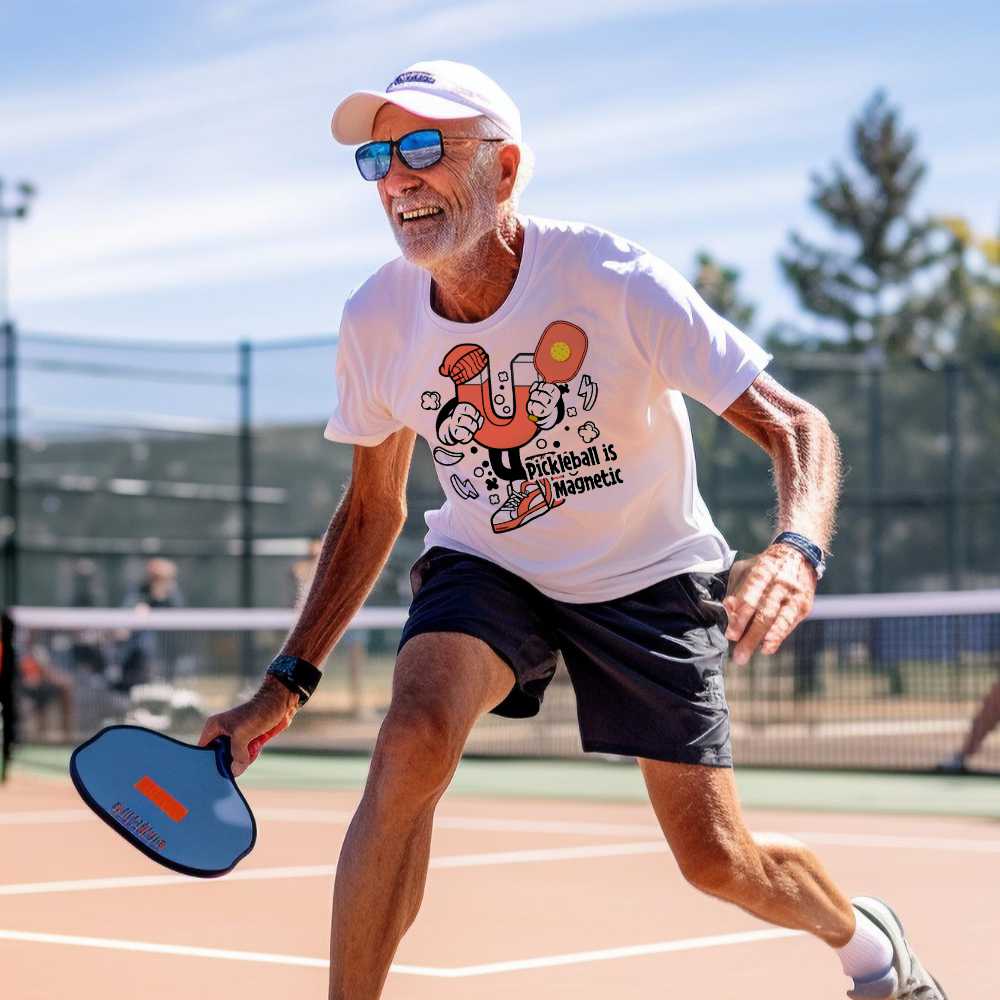 Senior pickleball player wearing a graphic white t-shirt with a humanoid magnet holding a pickleball paddle. The inscription states:"Pickleball is Magnetic."
